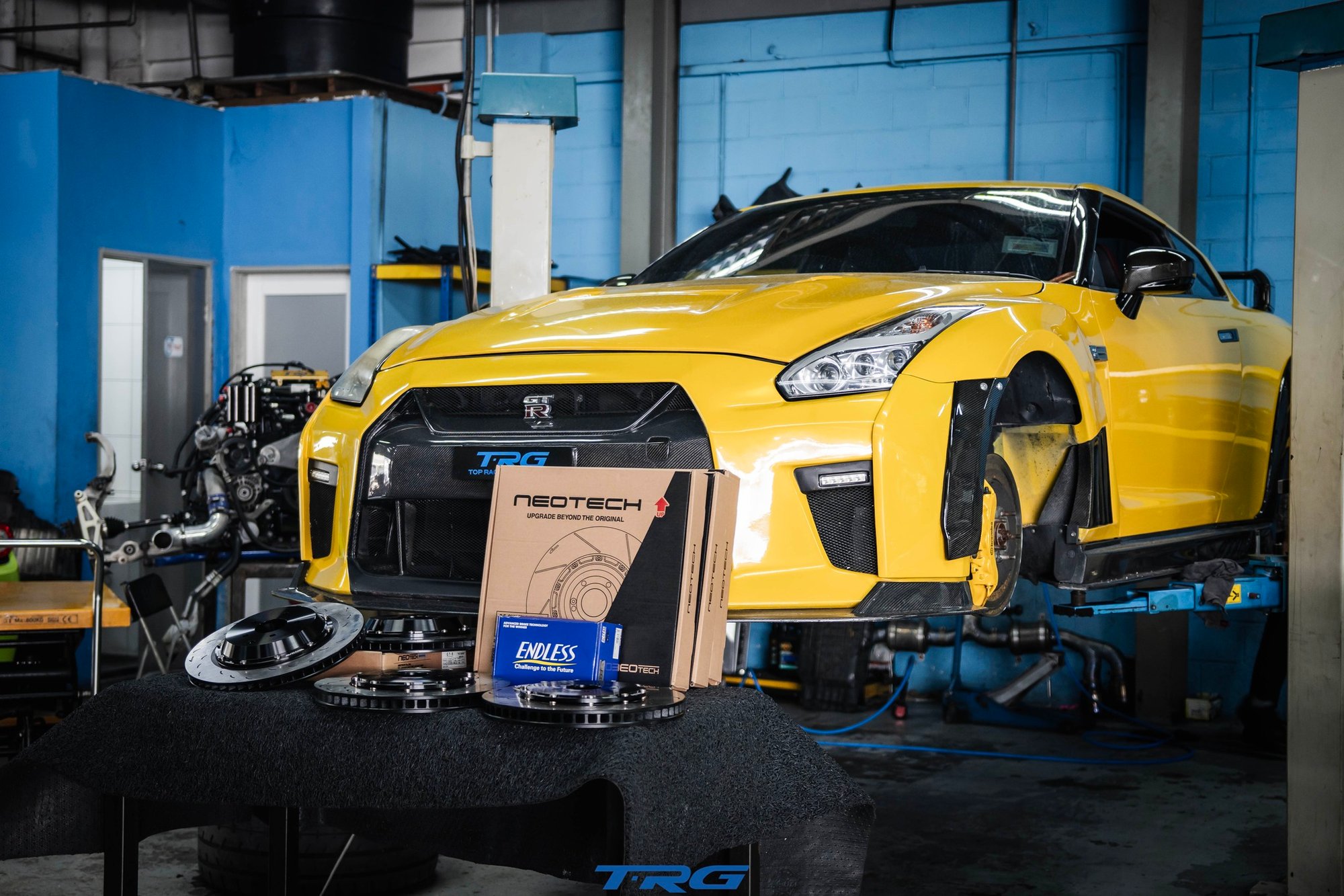Yellow widebody Nissan GT-R R35 on the TRG lift, wheels off, Neotech brake discs and cartons on the bench